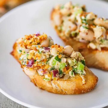 A close-up view of two pieces of toasted crostini that are both topped with an albacore tuna and white bean salad that has toasted bread crumbs on top .