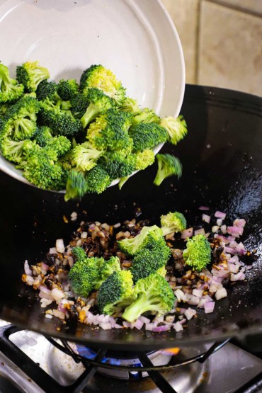 A person transferring broccoli florets from a white bowl into a wok on a gas stove with sautéed shallots, ginger, and garlic in it.