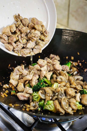 Chunks of cooked chicken thighs transferring from a white bowl into a wok on a gas stove that is filled with sautéed mushrooms, broccoli, and vegetables.