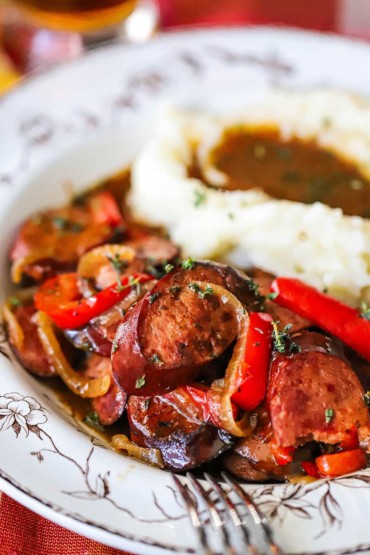 A close-up view of an antique bowl filled with a helping of beer-braised kielbasa and peppers next to a pile of mashed potatoes and gravy.