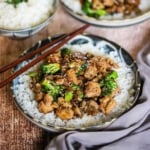 A close-up view of a shallow bowl filled with a layer of white rice topped with chicken and broccoli stir-fry with mushrooms.