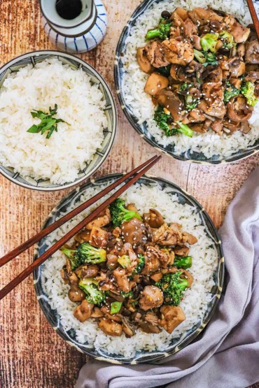 An overhead view of two shallow bowls filled with white rice topped with chicken and broccoli stir-fry both sitting next to a bowl of white rice.