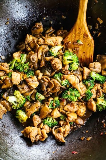 An overhead view of a wok filled with stir-fried chicken, broccoli, and mushrooms with a wooden spoon inserted off to the side.