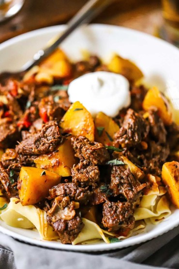 A close-up view of a white bowl filled with Hungarian goulash over egg noodles and topped with a dollop of sour cream.