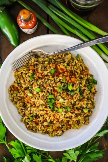 An overhead view of a white serving bowl filled with a pile of Cajun directy rice with a fork stuck on the side and fresh scallions and parsley along the outside of the bowl.