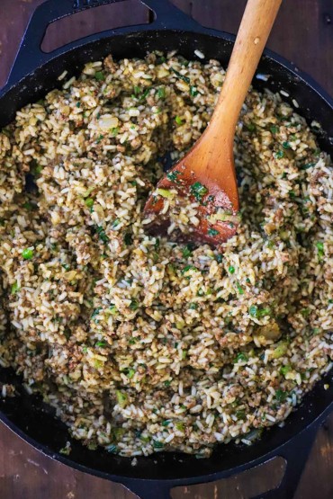 An overhead view of a large black cast-iron skillet filled with Cajun dirty rice with a big wooden spoon inserted into the middle of it.