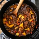 An overhead view of a black cast-iron pot filled with Hungarian goulash with chunks of beef, yellow potatoes, red bell peppers, and a brown sauce.