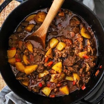 An overhead view of a black cast-iron pot filled with Hungarian goulash with chunks of beef, yellow potatoes, red bell peppers, and a brown sauce.