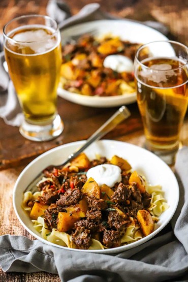 A straight-on view of two bowls of Hungarian goulash sitting next to two tall glasses of amber beer.