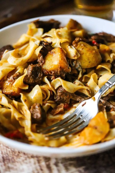 A close-up view of a white bowl that is filled with Hungarian goulash tossed with egg noodles and a portion of it missing.