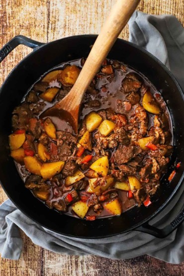 An overhead view of a black cast-iron pot filled with Hungarian goulash with chunks of beef, yellow potatoes, red bell peppers, and a brown sauce.