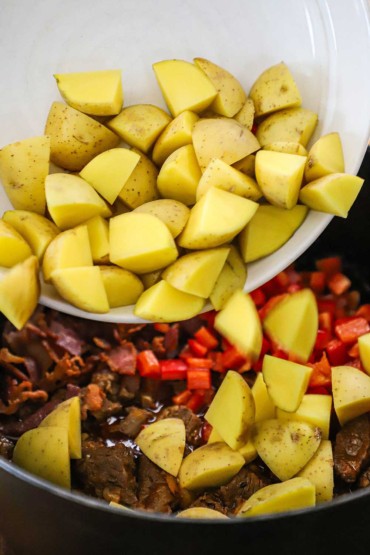 A person dumping quartered chunks of gold potatoes into a pot of simmering chunks of beef, bell peppers in a beef sauce.
