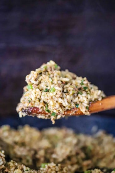 A large wooden spoon raising a heaping pile of Cajun dirty rice from a large black skillet filled with the same.