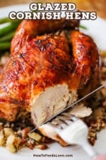 A person using a silver steak knife to cut away a slice of meat from the side of a glazed Cornish hen sitting on bed of wild rice on a white plate.