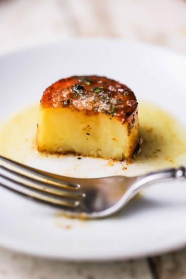 A front view of a half-eaten fondant potato sitting on a small white plate with a fork next to it.