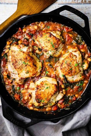 An overhead view of a large black cast-iron skillet filled with four pieces of Tuscan chicken with white beans and spinach.