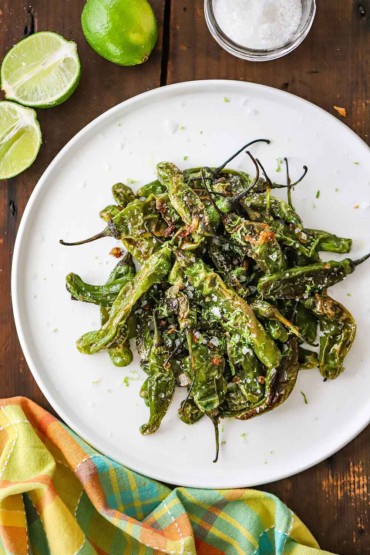 An overhead view of a white serving plate filled with a pile of seared shishito peppers all next to a couple of limes and a small jar of sea salt.