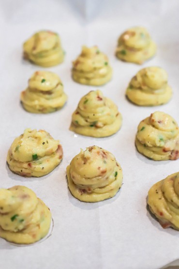 Several rows of bacon parmesan gougères mounds of dough on a parchment paper on a baking sheet.