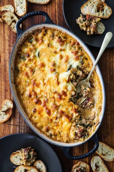 An overhead view of an oval baking dish filled with a partially eaten sausage, bean, and spinach dip with a silver spoon inserted into the side of the dip.
