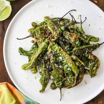 An overhead view of a white serving plate filled with a pile of seared shishito peppers all next to a couple of limes and a small jar of sea salt.