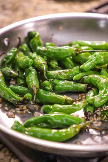A large stainless steel skillet filled with shishito peppers and chopped shallots that are being sautéed.