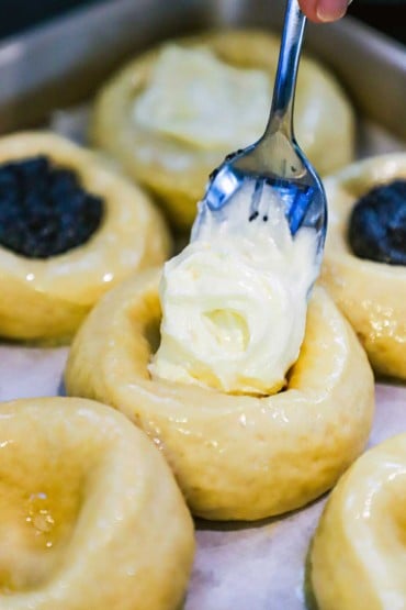 A person using a spoon to transfer a mound of cream cheese filling into a well of an uncooked kolache pastry.