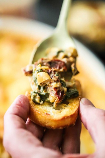 A person holding a toasted crostini with one hand while the other hand is spooning a sausage, bean, and spinach dip onto the crostini.