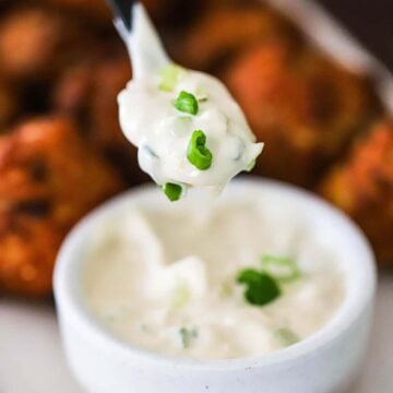 A small spoon being held up holding a spoonful of white remoulade topped with chopped scallions over a bowl of the same.