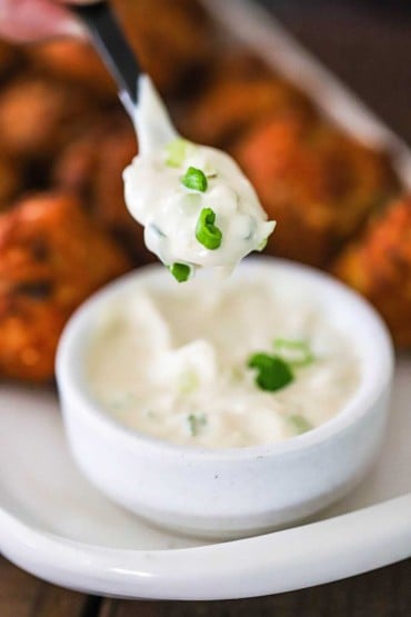 A small spoon being held up holding a spoonful of white remoulade topped with chopped scallions over a bowl of the same.