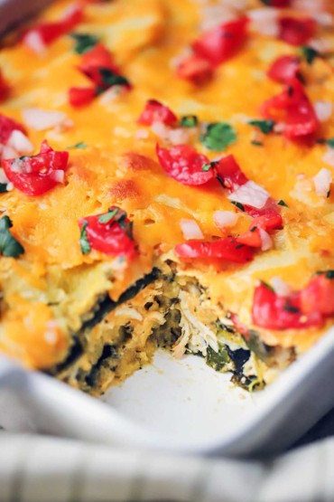 A close-up view of a green enchilada casserole with a serving missing in a white dish with pico de gallo sprinkled on top.