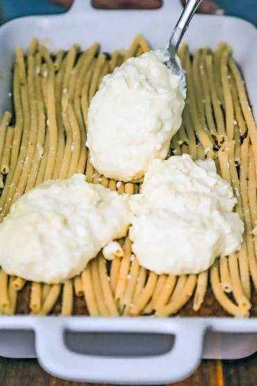 A person spooning a large amount of thick Greek bechamel sauce over tubular pasta on meat sauce in a white lasagna dish.