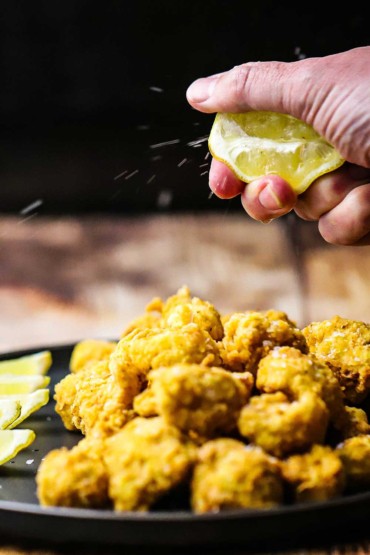 A person squeezing a lemon wedge over black circular plate filled with fried cauliflower and lemon wedges.
