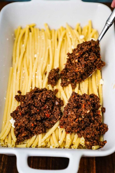 A person transferring a pastitsio meat sauce onto Greek tubular pasta in a large white lasagna dish.