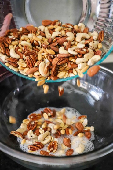 A person pouring a variety of nuts from one large glass bowl into another large glass bowl with foamy egg whites in it.