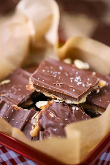 A close-up view of a pile of chocolate toffee with almond and sea salt in a festive red and white checkered Christmas tin.