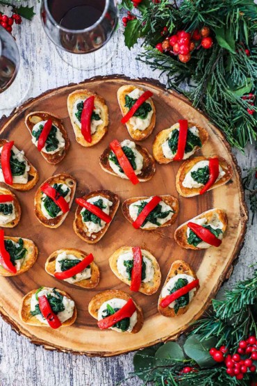 An overhead view of a wooden circular platter filled with toasted crostinis topped with a white bean spread, spinach, and roasted red bell pepper.