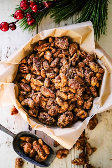 An overhead view of spiced nuts that are in a large tin that is lined with brown paper and a scooper next to it filled with more of the spiced nuts.