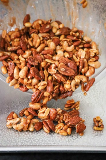 An assortment of nuts that have been coated in egg whites and spiced sugar being dumped from a glass bowl onto a large baking sheet.