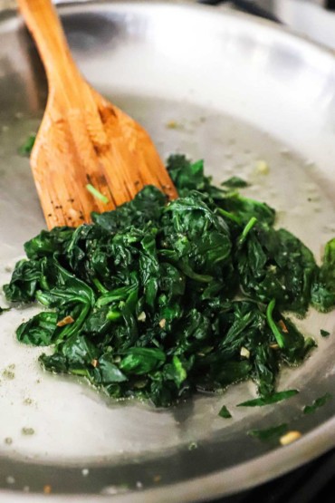 A large silver skillet filled with wilted sautéed spinach with a wooden spatula off to the side.