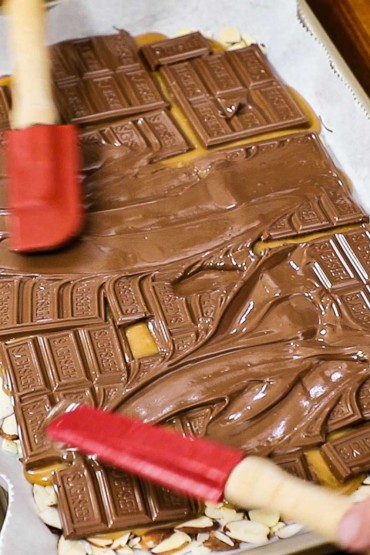 Two small red spatulas being used to smear Hershey chocolate bars that are partially melting over a layer of warm toffee on top of almond slivers.