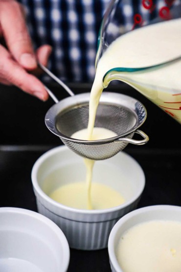 A person pouring a thin custard through a small sieve into a small white ramekin.