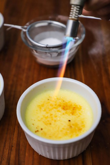A person using a kitchen blow torch to caramelize sugar on top of a small white ramekin filled with crème brûlée.