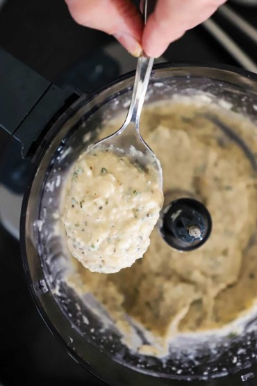 A person holding up a spoon filled with a white bean purée over a food processor.