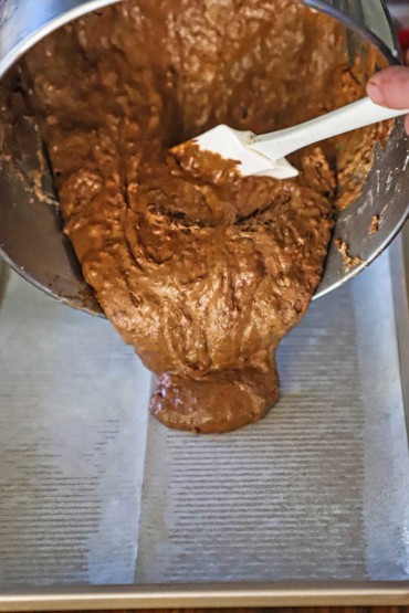 A person using a white spatula to transfer a chocolate cake batter from a mixing bowl onto a baking sheet that is lined with oiled parchment paper.
