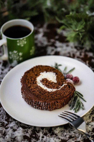 A slice of a yule log sitting on a white dessert plate next to a festive holiday mug filled with black coffee.