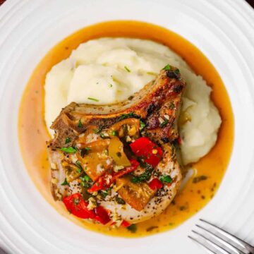 An overhead view of an Italian-style smothered pork chop resting on a bed of mashed potatoes with a pool of sauce in the bottom of the plate.