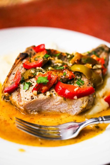 A close-up view of a partially eaten pork chop that is covered with Italian peppers and herbs and resting in an amber-colored sauce.