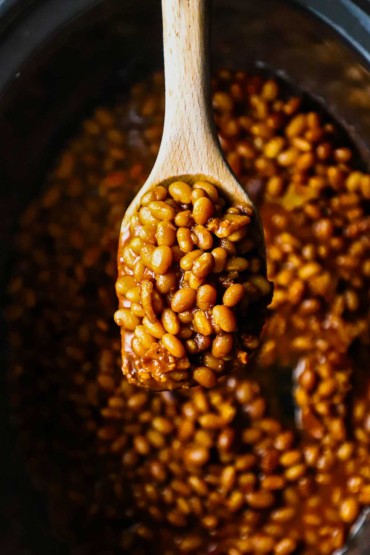 An overhead view of a large wooden spoon holding a big helping of slow cooker baked beans over the slow cooker pot filled with the same.