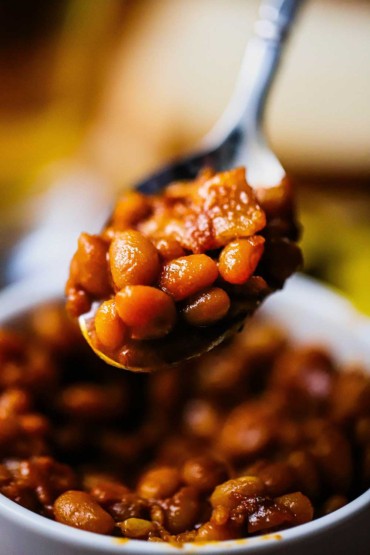 A silver spoon being held up that is holding a helping of slow cooker baked beans over a small bowl containing a full serving of the beans.