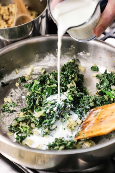 A person pouring whole milk from a glass measuring cup into a skillet that is filled with sautéed spinach, onion, and garlic.